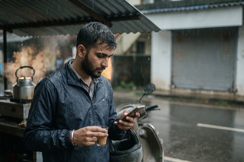 A man at a roadside tea stall in the rain, answering a customer on his phone between visits