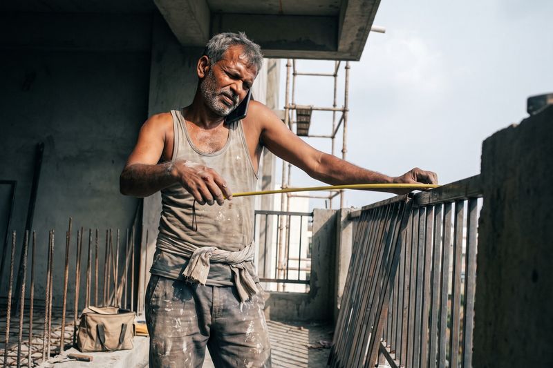 A fabricator measuring a balcony grill, his phone held between ear and shoulder